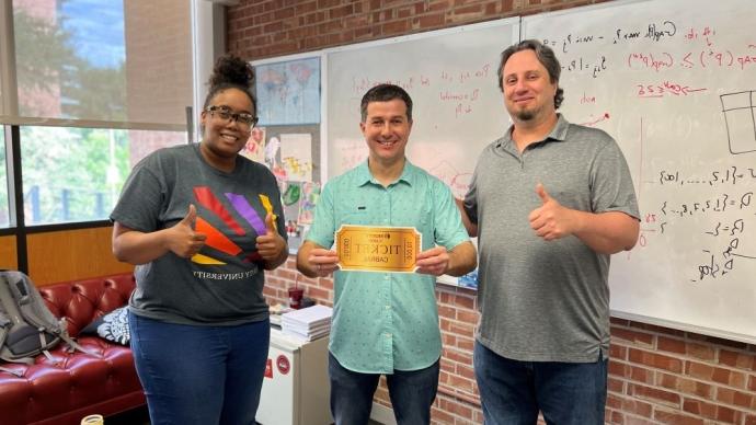 Three people standing with the man in center holding a golden ticket