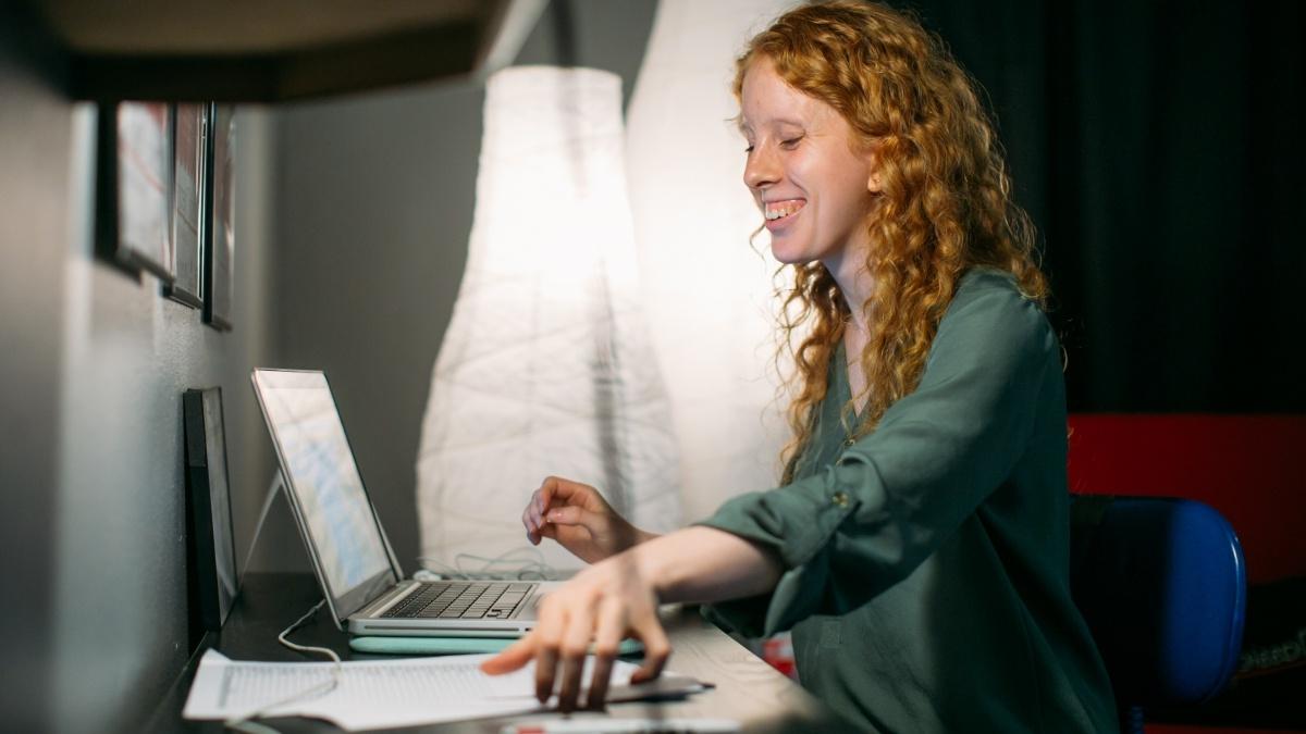 red headed person smiling while looking at a screen and grabbing for papers